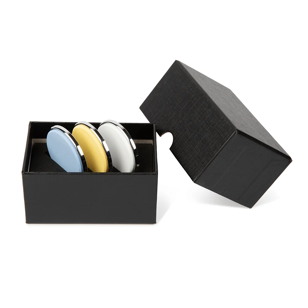 Un coffret cadeau noir avec couvercle amovible contient le Lot de 3 disques aiguiseur professionnel de Maison Damas - bleu, jaune et blanc, de forme ovale avec des bords chromés réfléchissants - joliment disposés les uns à côté des autres.